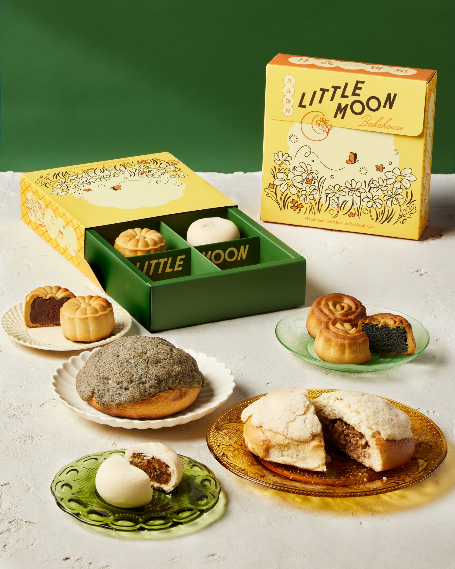 Little Moon Bakehouse Product Photo Group Shot with Gift Boxes | Photo by Dana Mariko Chang. Two yellow Little Moon Bakehouse gift boxes, one standing up and the other open on the table with snacks inside. Five plates of pastries laid out in the foreground with two breads, two mooncakes, and one Taiwanese pineapple cake. Background is green. Table is white.