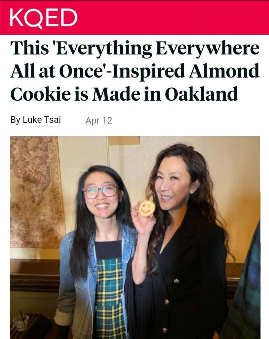 Annie Wang, owner of Little Moon Bakehouse, alongside Michelle Yeoh at the premiere of Everything Everywhere All At Once in San Francisco's Castro Theater. Michelle Yeoh holding Annie's T Cakes custom smile almond cookie. Both smiling at the camera.