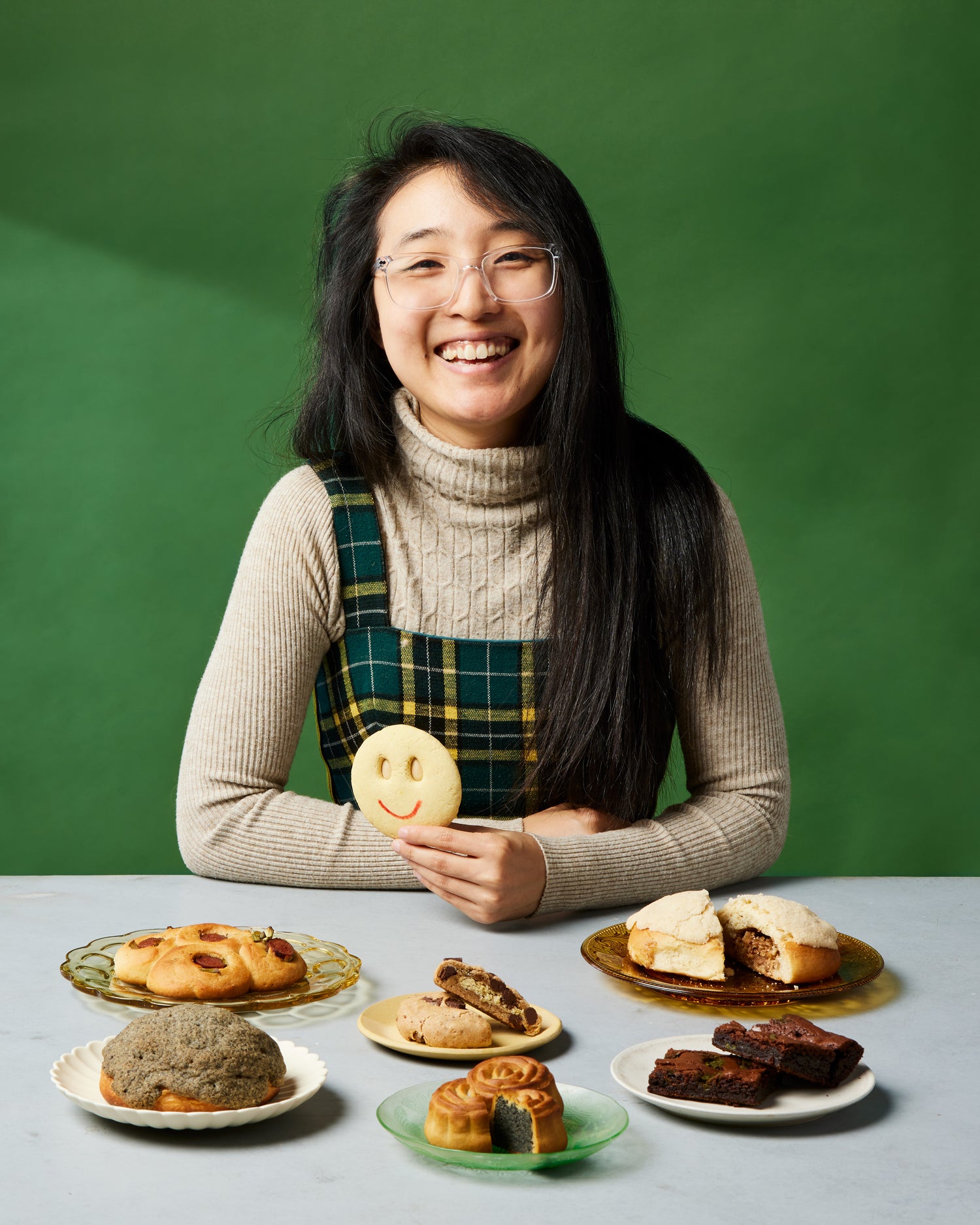 Headshot for Annie Wang of Little Moon Bakehouse | Photo by Dana Mariko Chang. Annie in a tan long sleeve sweater and a checkered yellow, green and black jumper sits and smiles in front of a green background. In front of her are plates of pastries she's made including three bread buns, one mooncake, one plate of mooncookies, and one plate of brownies. Annie is holding one of her signature smiley face almond cookies.