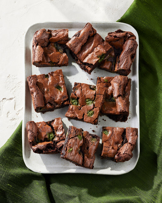 A white tray with nine dark chocolate brownies with matcha chocolate chunks throughout the brownies. The tray is on a half white half green cloth background. Gluten free.