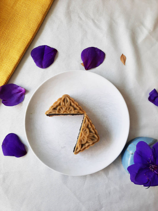 Overhead shot of black sesame mooncake cut in half. Background is white with purple flowers and brown leaves on a thin branch.